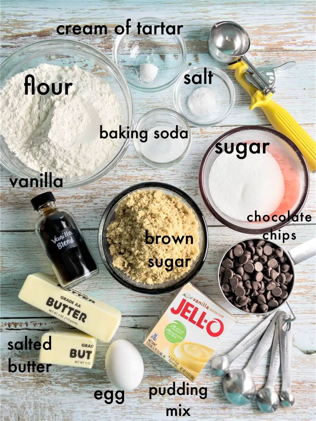 An overhead shot of chocolate chip cookie ingredients, labeled "brown sugar, sugar, chocolate chips, Jello instant pudding mix, salted butter, vanilla, flour, baking soda, salt, egg, and cream of tartar.
