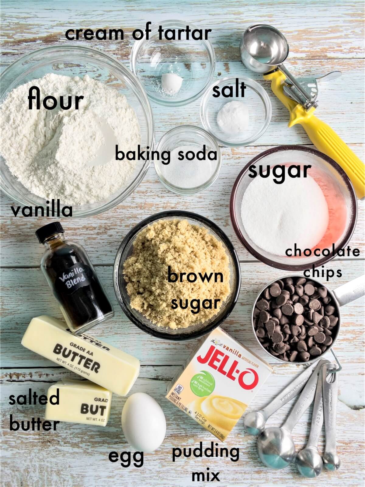 An overhead shot of chocolate chip cookie ingredients, labeled "brown sugar, sugar, chocolate chips, Jello instant pudding mix, salted butter, vanilla, flour, baking soda, salt, egg, and cream of tartar.