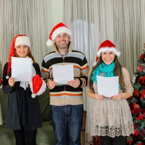 Three people with Santa hats on singing with sheet music for a Caroling Party theme idea..