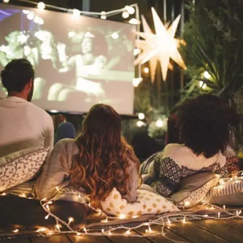 Several people watching a movie on a projector screen with Christmas lights around.