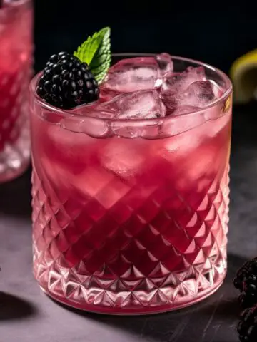 A pretty magenta blackberry gin cocktail with ice in a cocktail glass with a sprig of mint.
