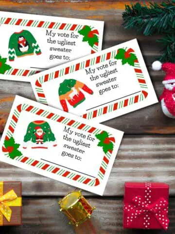 Three ugly christmas sweater voting ballots with the text "my vote for the ugliest sweater goes to:"on a wooden table with christmas decorations.