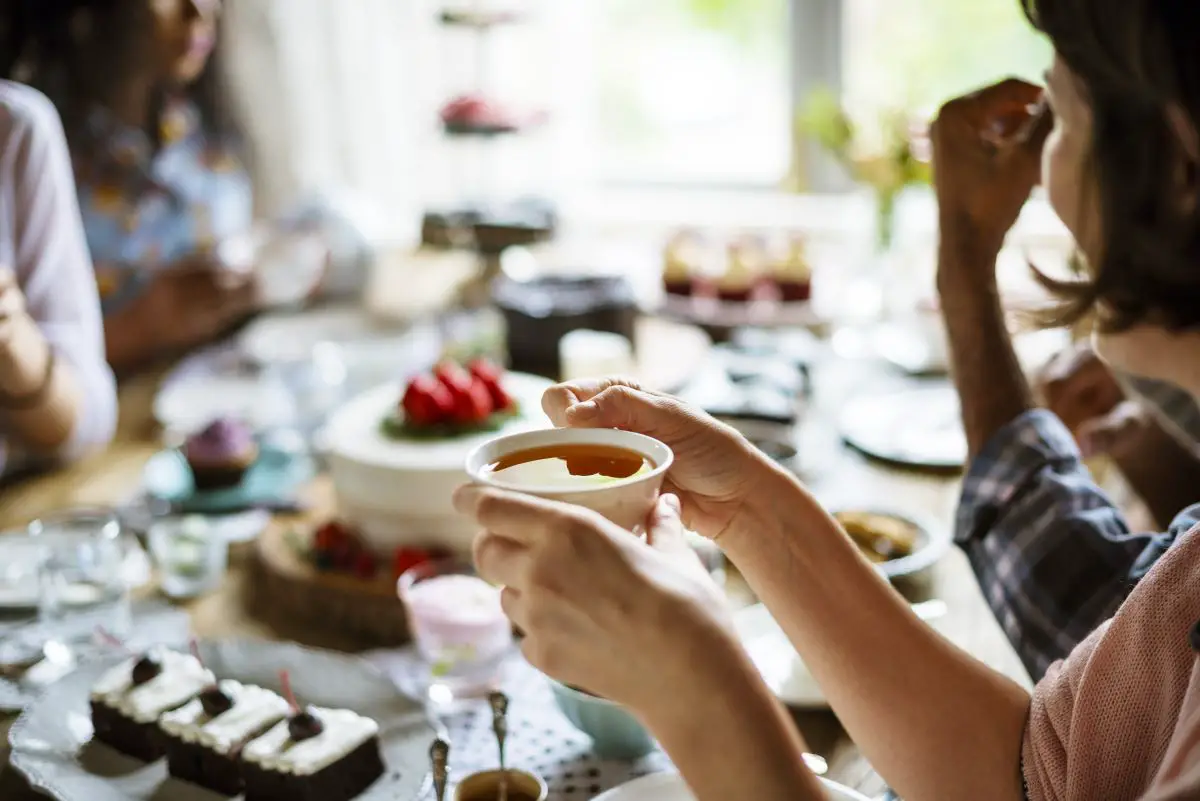 A woman and other adults around a table enjoying tea and crumpets and other treats.