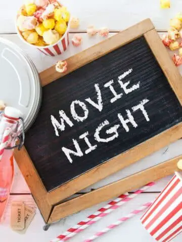A chalkboard that says "movie night" with striped movie-themed straws and cups with popcorn and bottles of pink lemonade.