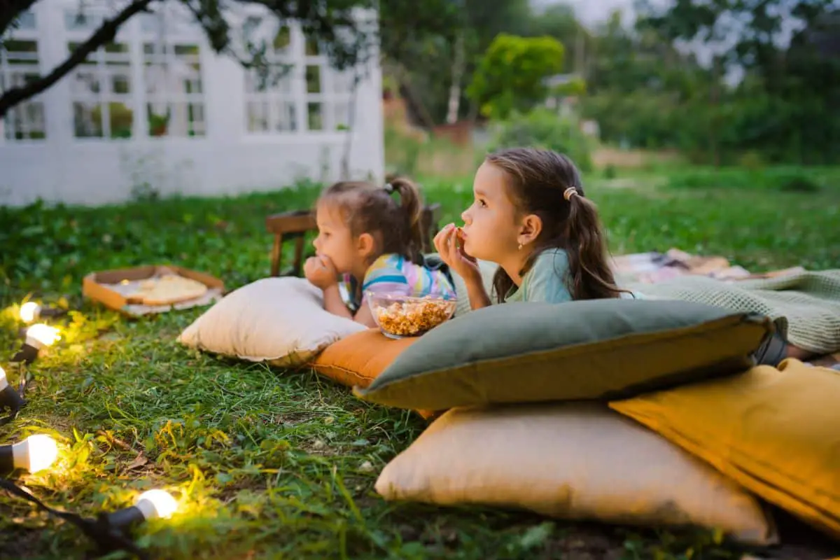 Two kids on pillows outside watching a movie.